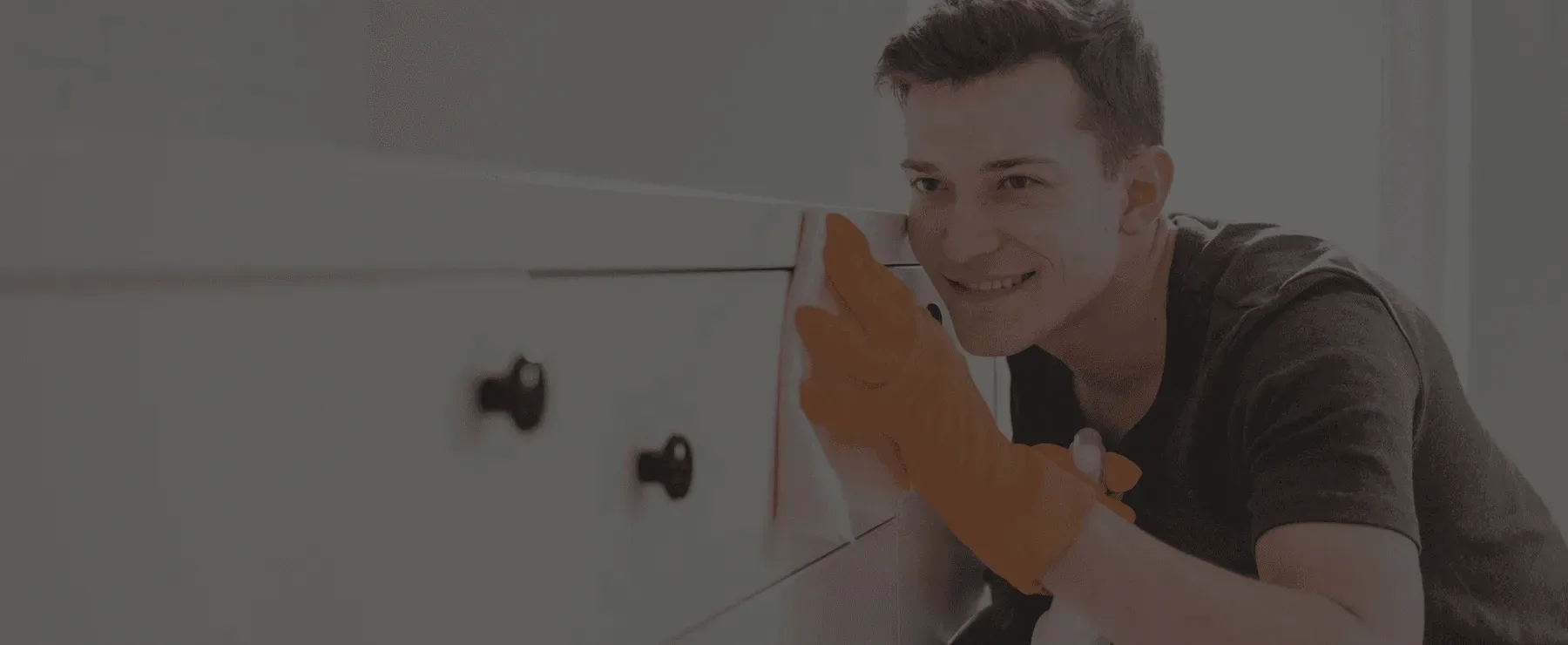 A smiling cleaner in orange gloves wiping a cabinet, under a dark veil.