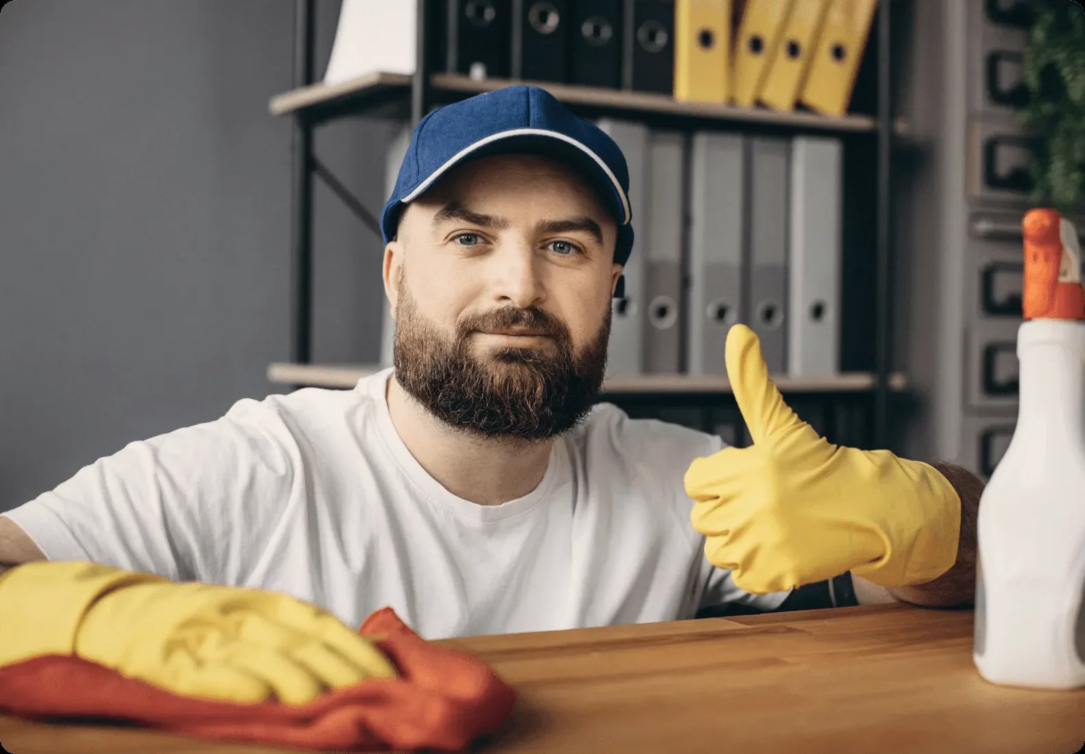 A bearded cleaner in a blue cap giving a thumbs up at a desk.