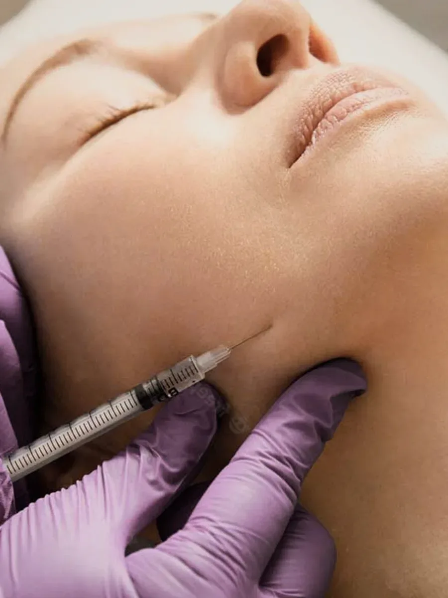 A face under a syringe held by a purple-gloved hand.