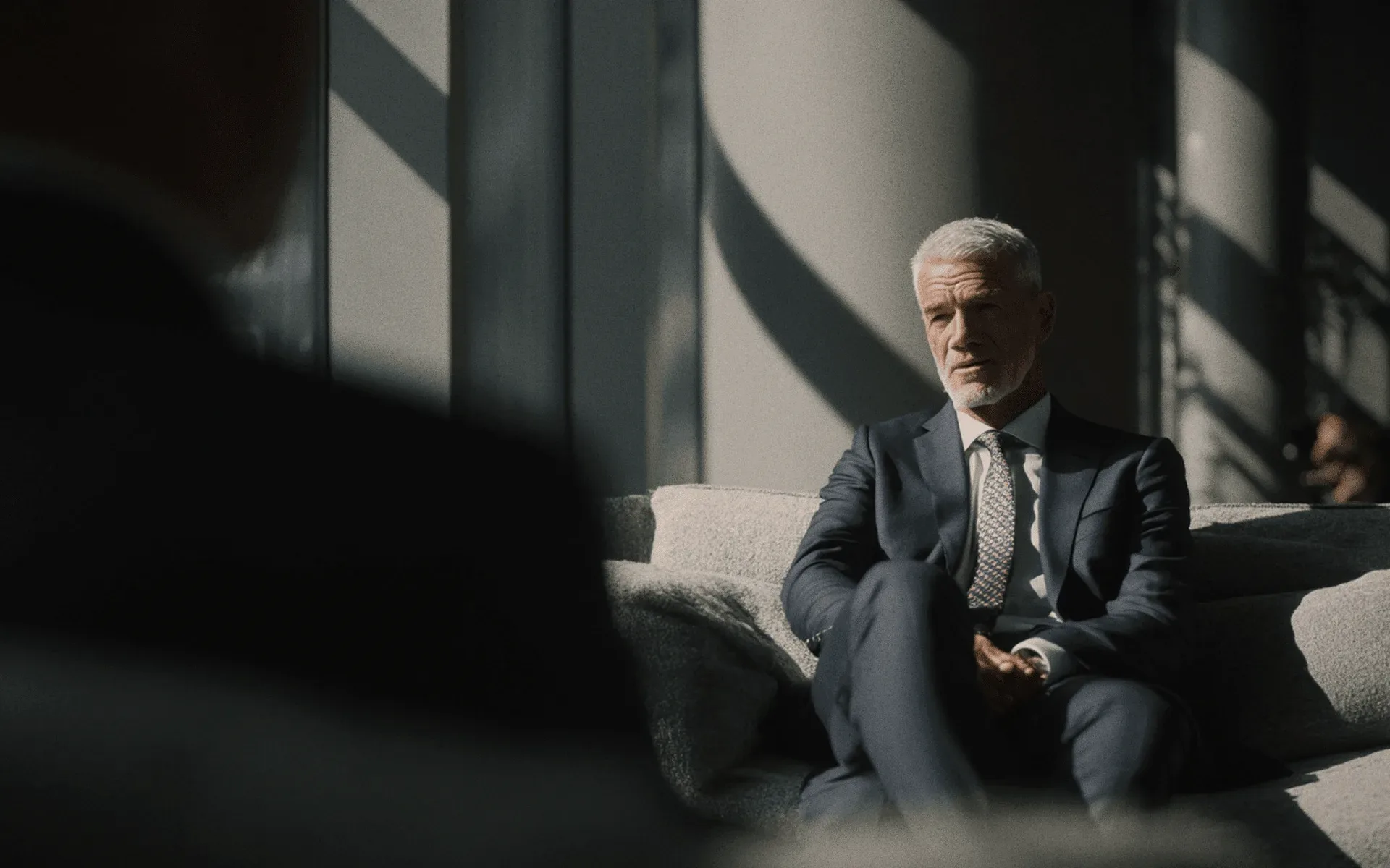A man in a navy suit on a grey sofa in striped light.