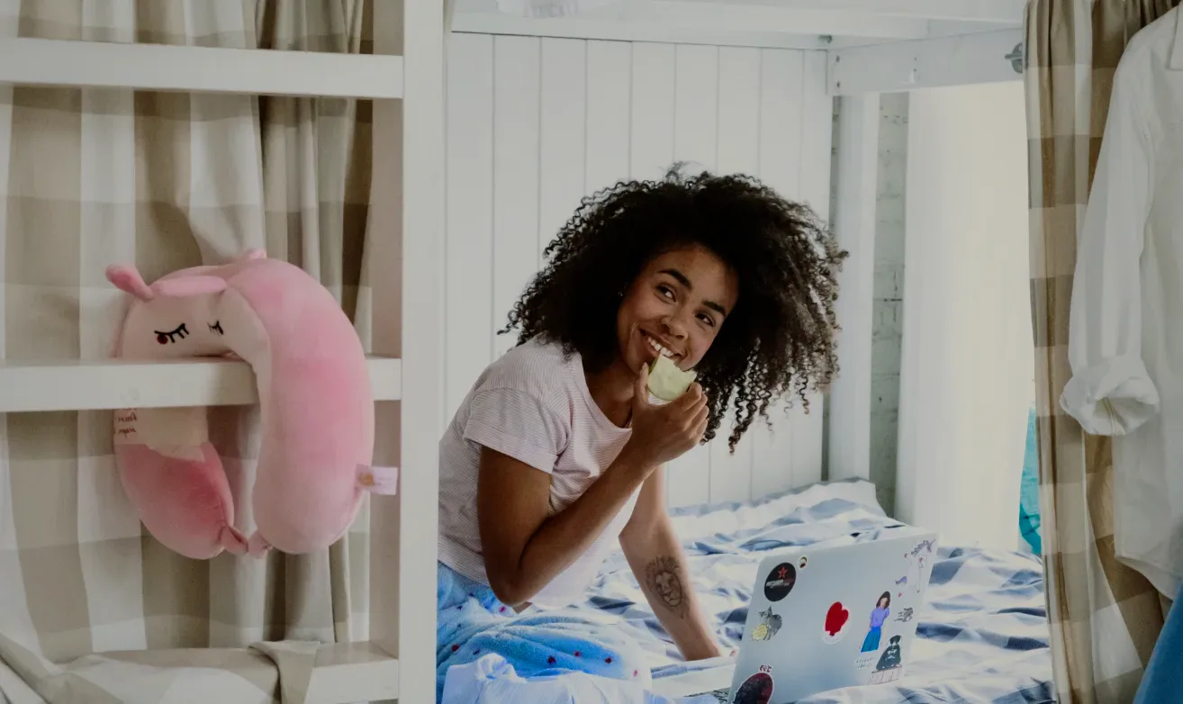 A guest eating an apple on a bunk bed beside her laptop.