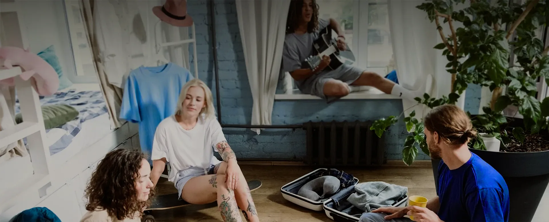 Young travellers relaxing in a bright hostel room, one playing guitar on the window sill.