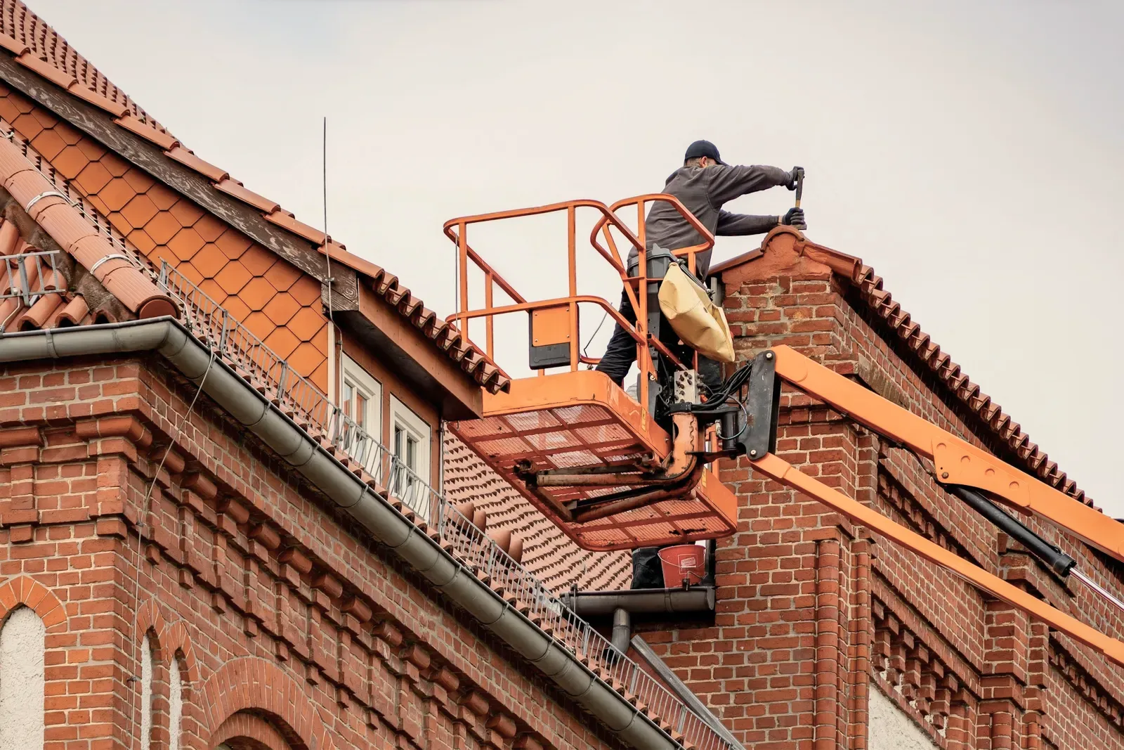 A worker on a raised platform working at the eaves of a tiled roof on a brick building.
