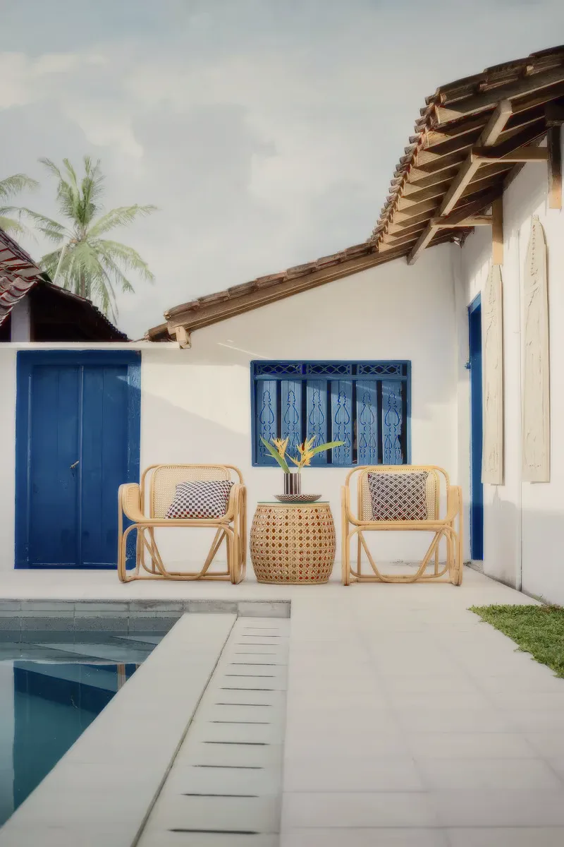 Two rattan chairs on a white patio beside a pool, blue doors behind.