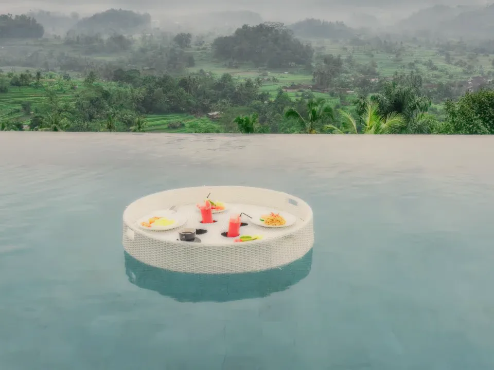 A floating breakfast tray in an infinity pool above green hills.