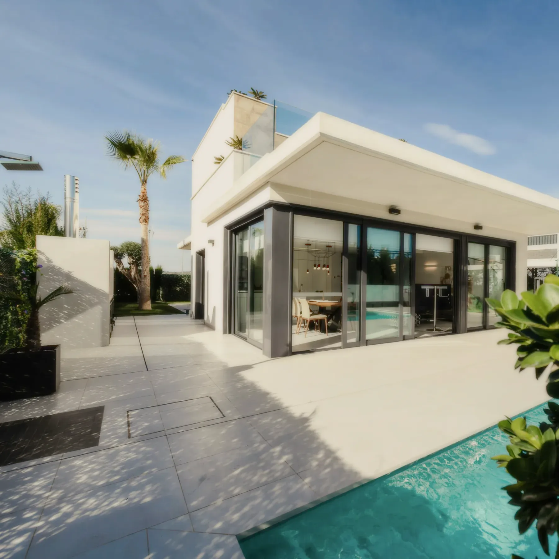 A white modern house with glass doors opening onto a stone terrace and a pool, a palm tree against a blue sky.