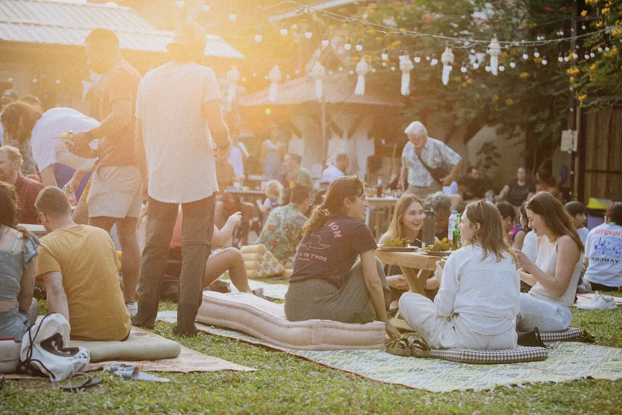 Guests on floor cushions in a garden at golden hour, under strings of white Lanna lanterns.