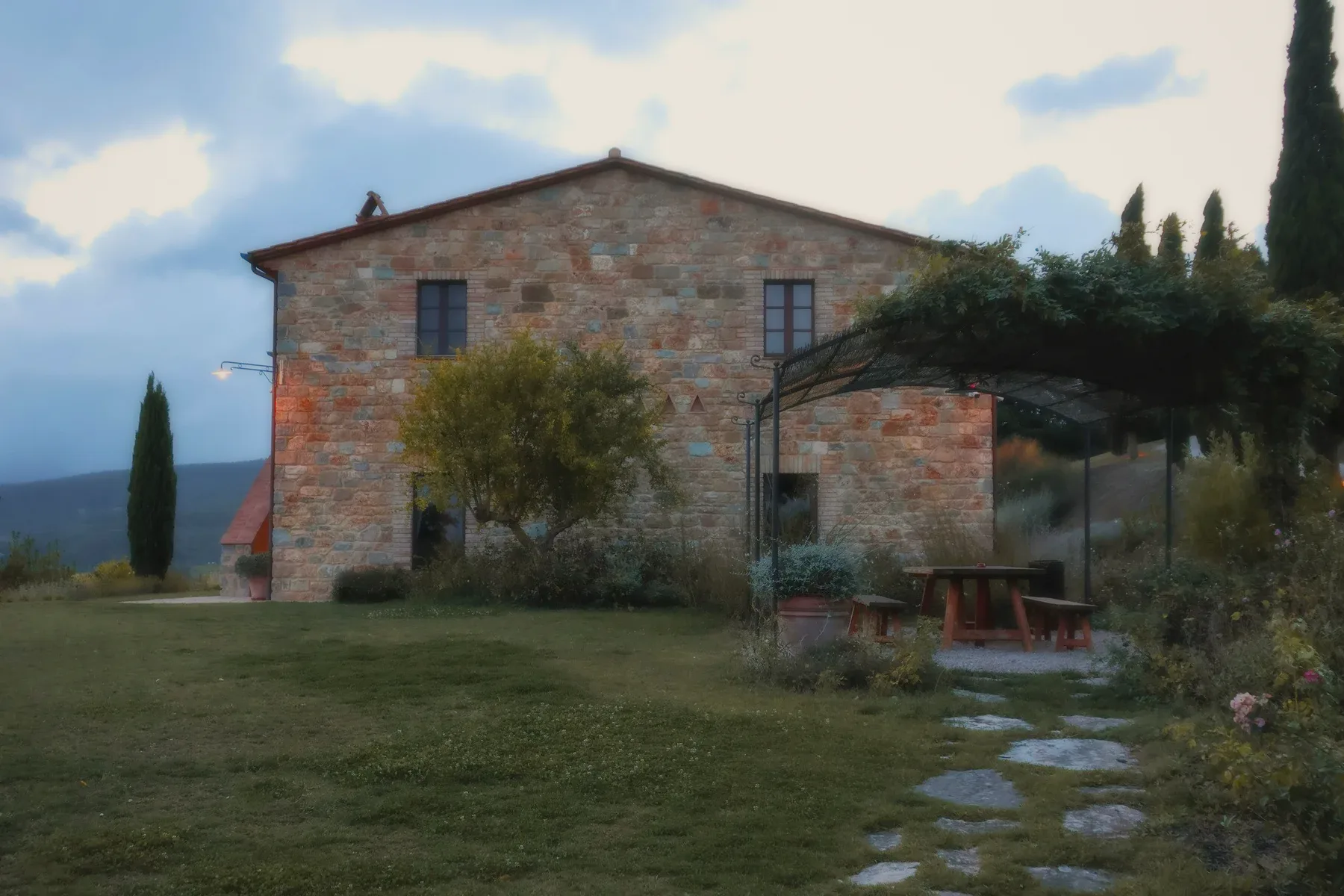 A stone villa at dusk, a pergola and a table on the lawn.