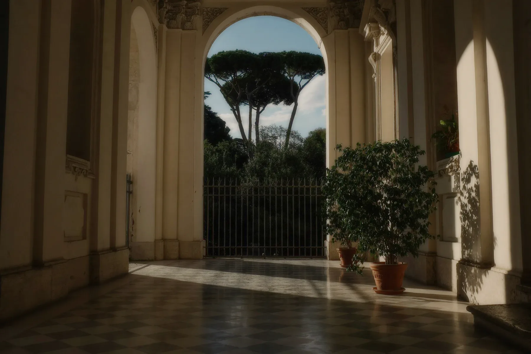 A stone arcade with potted trees, a gate and pines beyond.