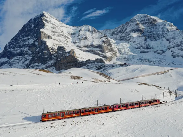 A red train crossing the snow below high peaks.