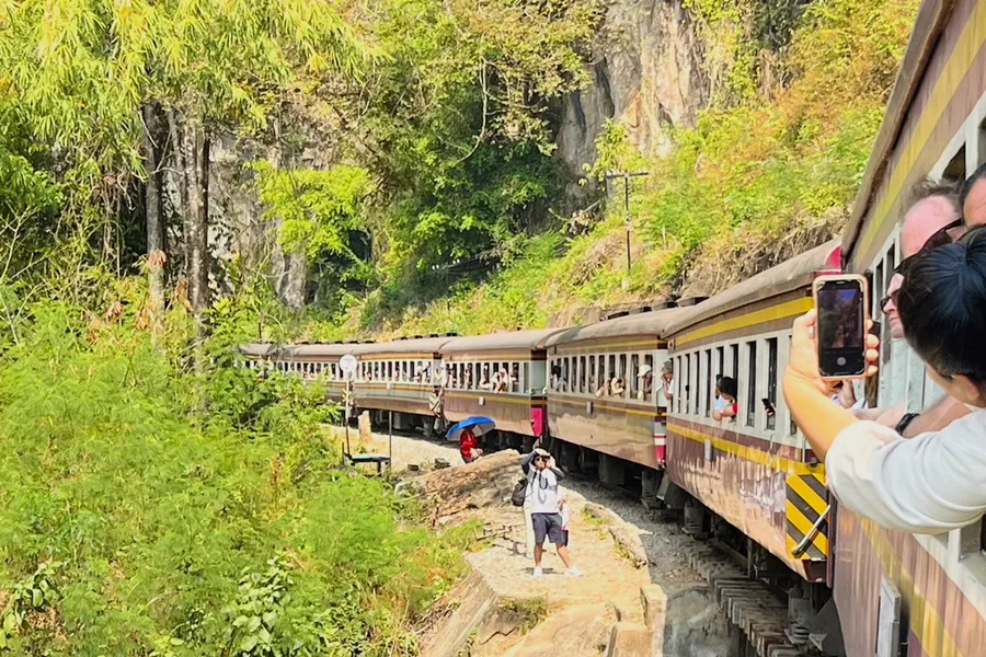 A train on a wooden viaduct beside a cliff, with a traveller taking a photograph.