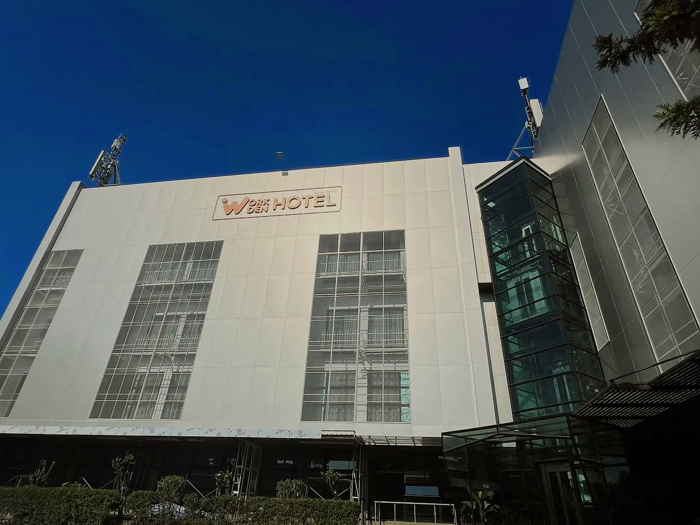 Work Den Hotel's white front with its name high on the wall, under a deep blue sky.