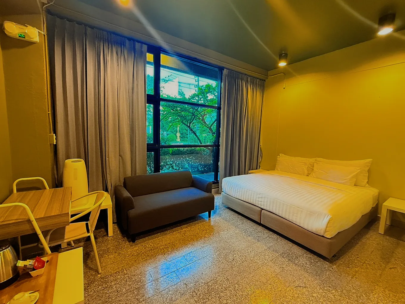 A double room with a yellow wall, a sofa and a tall window onto trees.