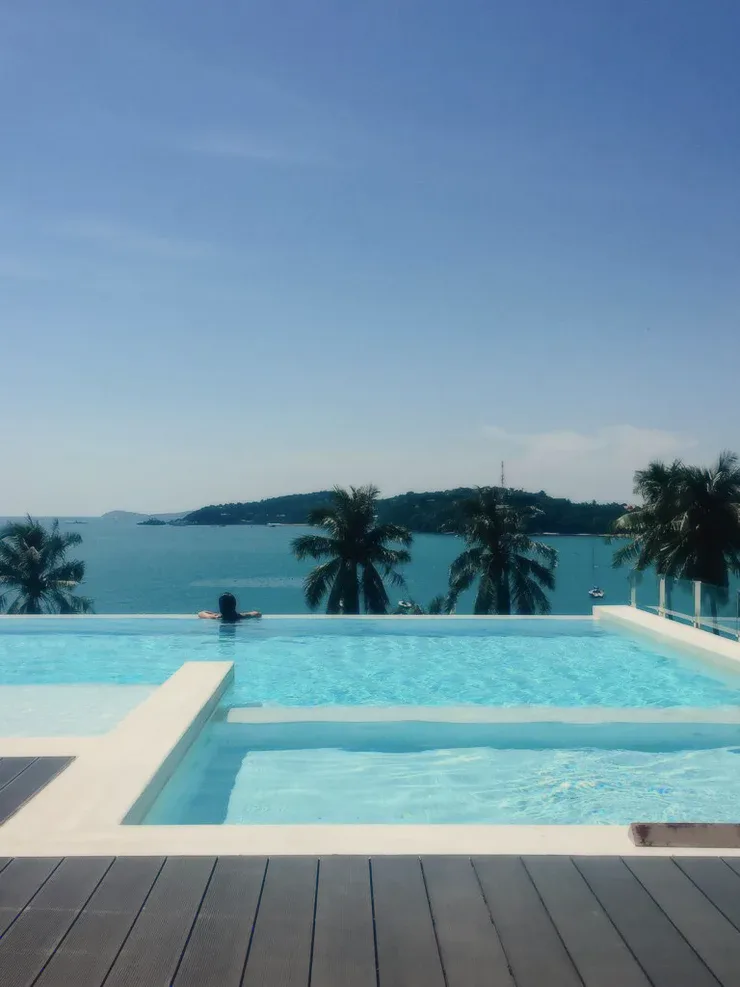 A pool on a wooden deck above a bay with palm trees.