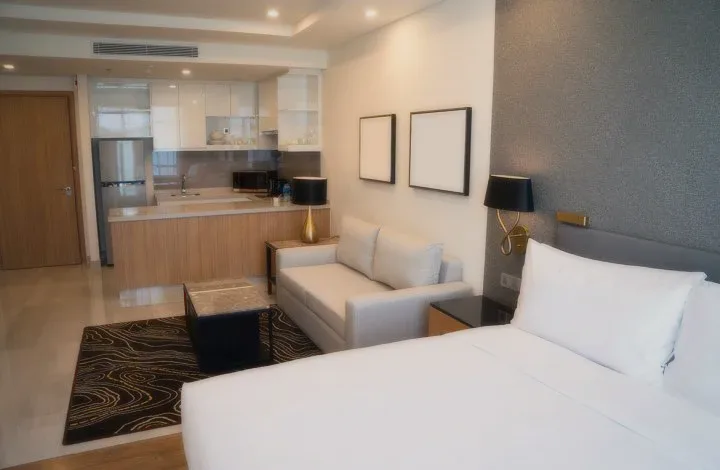 A bedroom with a king bed, a lounge chair and a kitchenette behind.